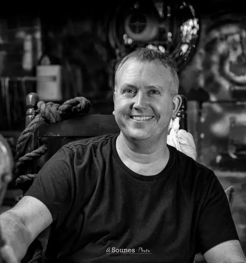 Black-and-white portrait of Matt McConnell seated in a rustic workshop, surrounded by sculptural elements