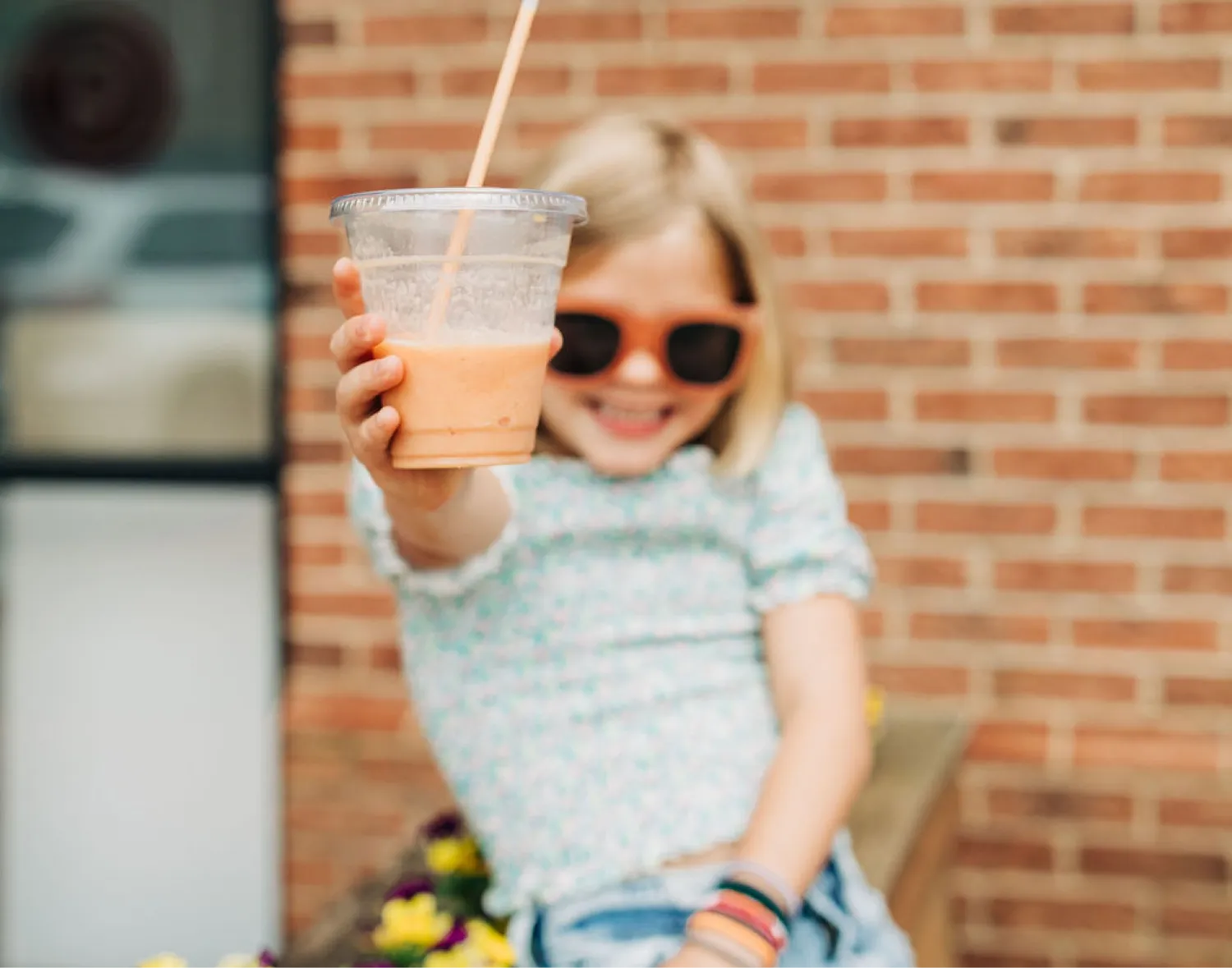 Smiling child wearing sunglasses posing in front of a brick café backdrop holds a fruit smoothie